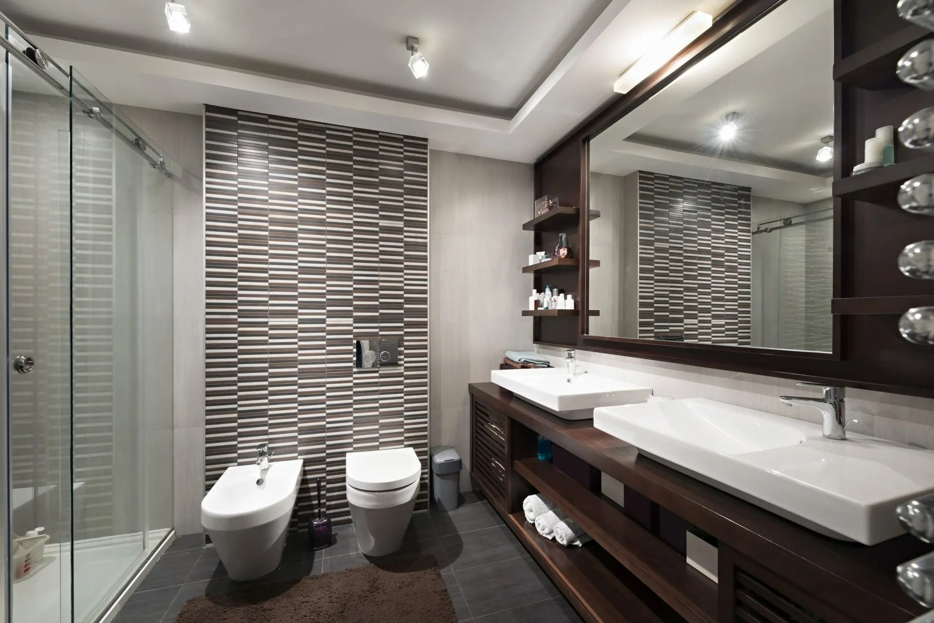Salle de bains moderne avec douche en verre, deux lavabos et toilettes. Couleurs neutres et murs carrelés.