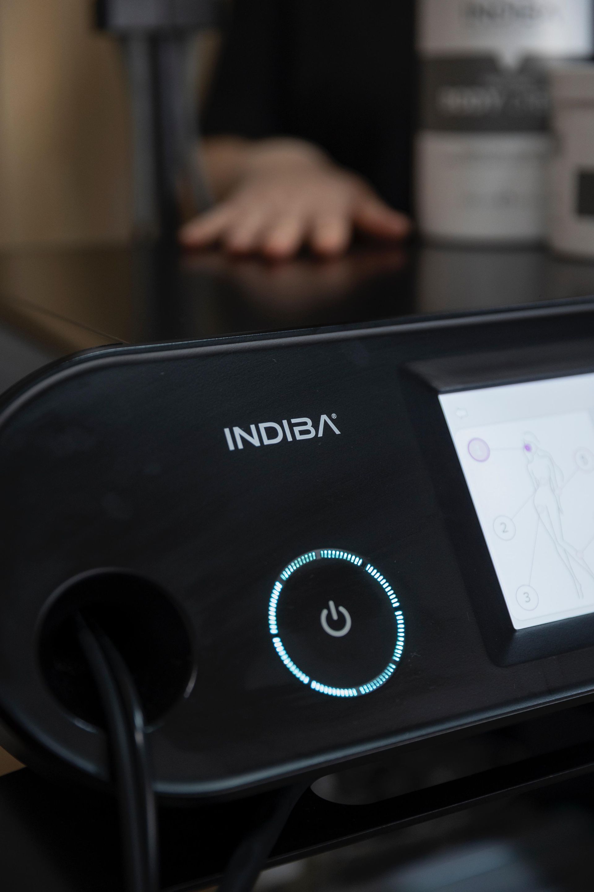 Dispositivo INDICA negro con botón de encendido brillante sobre una mesa. Una mano se ve borrosa al fondo.