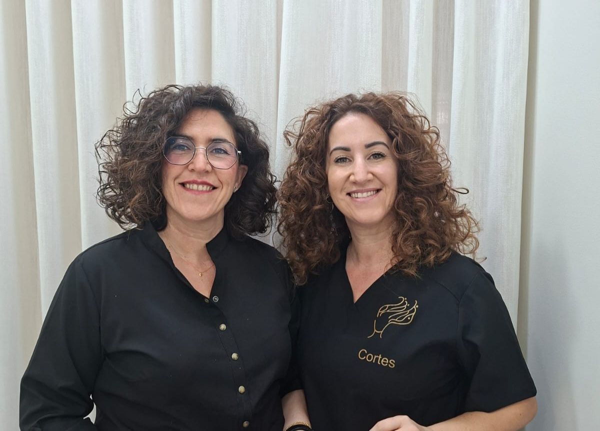 Dos mujeres de pelo rizado sonríen frente a una cortina. Una lleva gafas y camisa negra. La otra lleva un logo en la camisa.