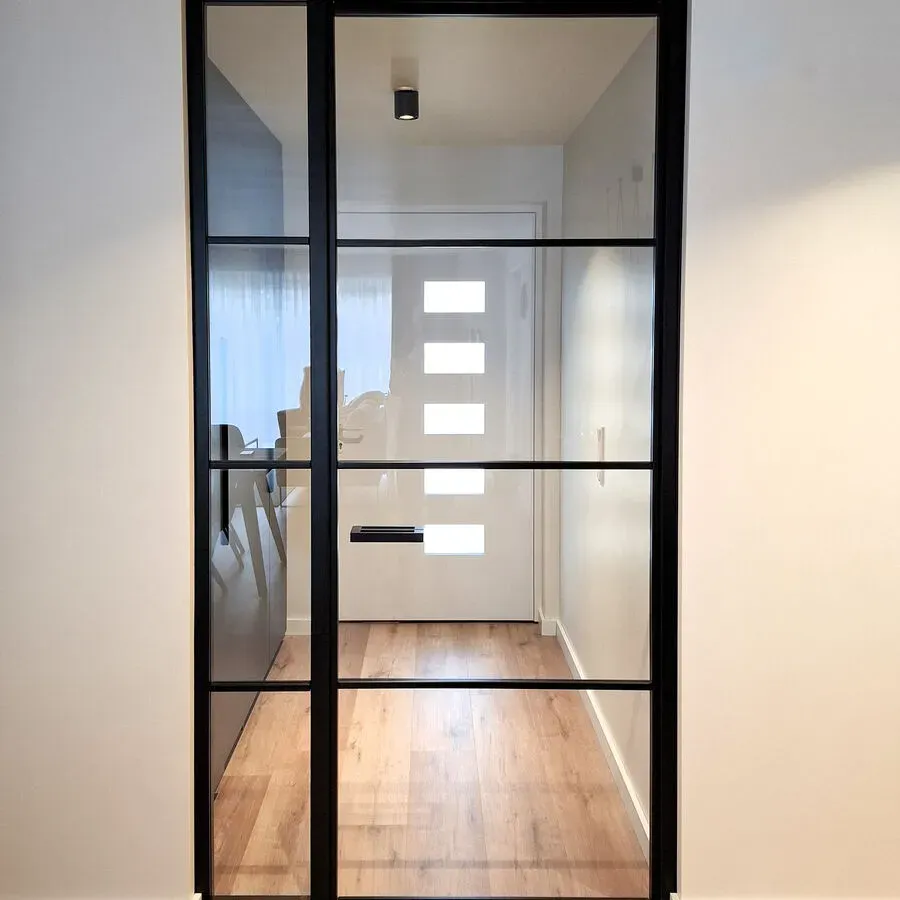 Schwarze Glastür mit Blick auf den Flur. Der Innenraum ist mit hellen Holzböden und weißen Wänden ausgestattet.