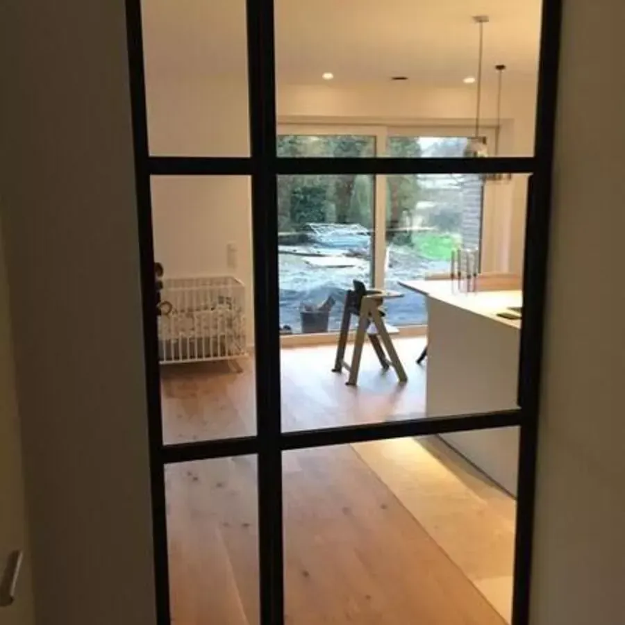 Schwarze Glastür, die den Blick ins Innere mit Kinderbett und Küche freigibt, sowie nach außen hin auf die Baumaterialien.