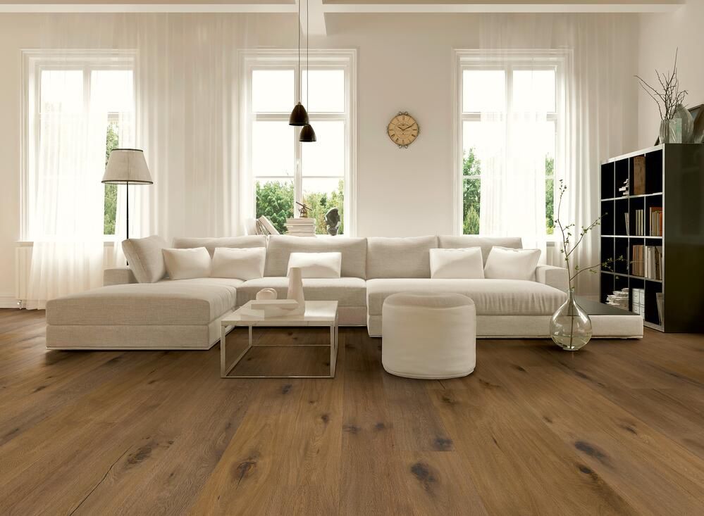 Wohnzimmer mit hellem Hartholzboden, großem Sofa, Hocker und Fenstern mit weißen Vorhängen.