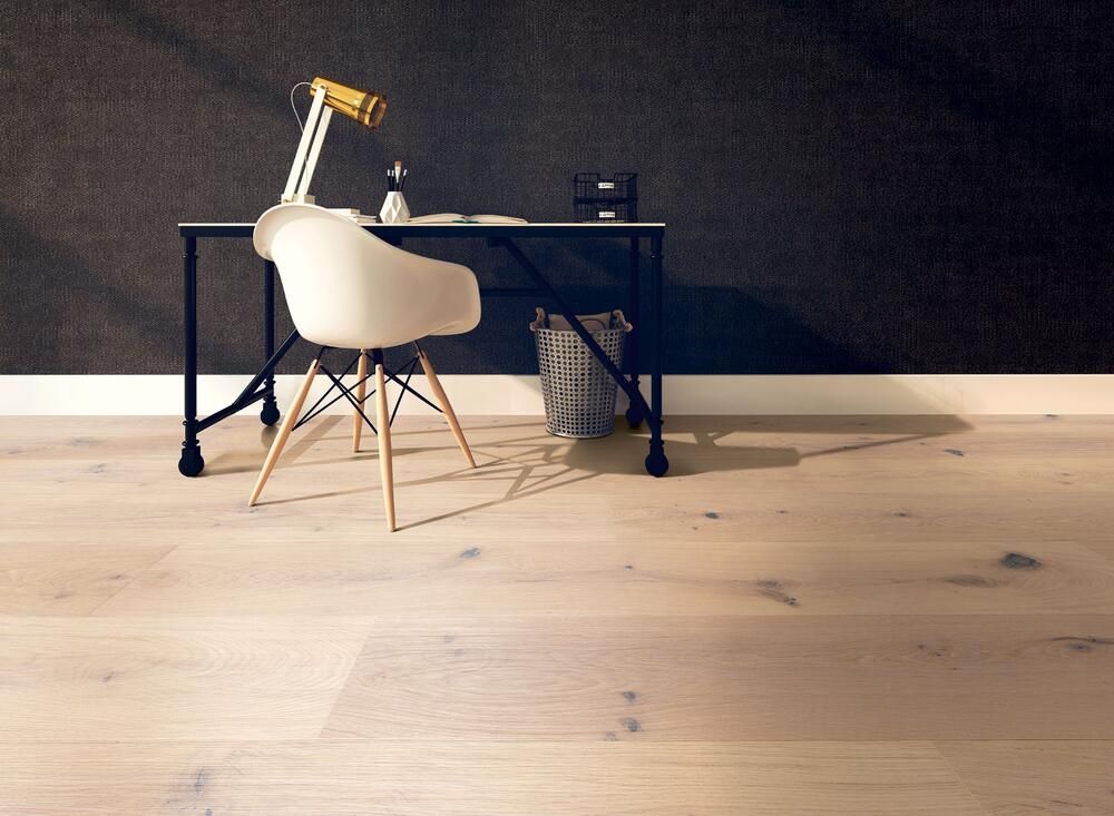 Schreibtisch mit weißem Stuhl, Lampe und Abfalleimer auf hellem Holzboden vor einer dunklen Wand.