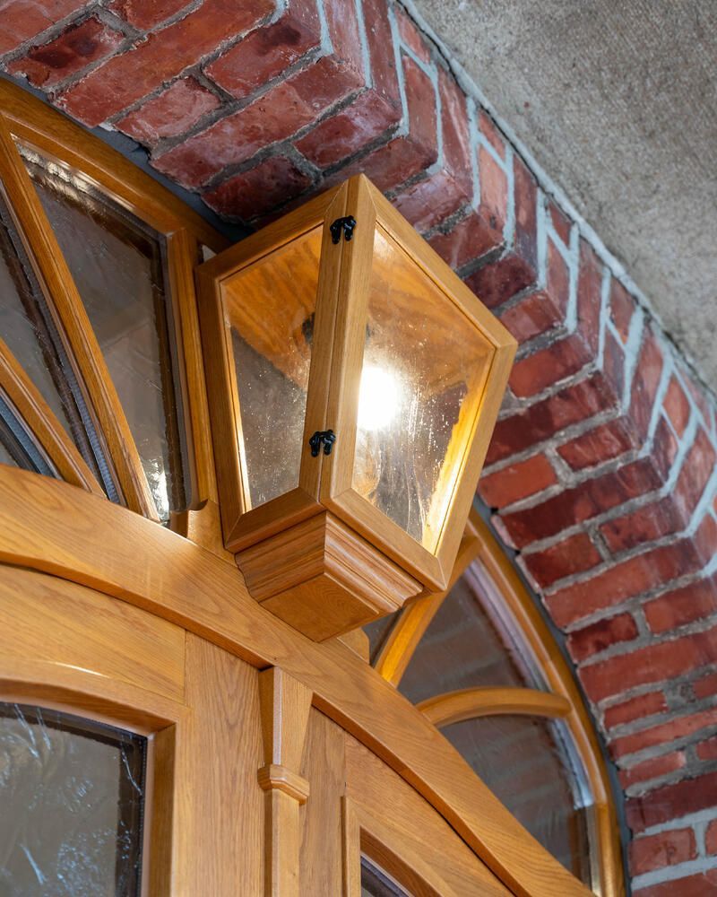 Eine hölzerne Laterne mit Glasscheiben ist über einer Holztür angebracht, vor einem roten Backsteinbogen.