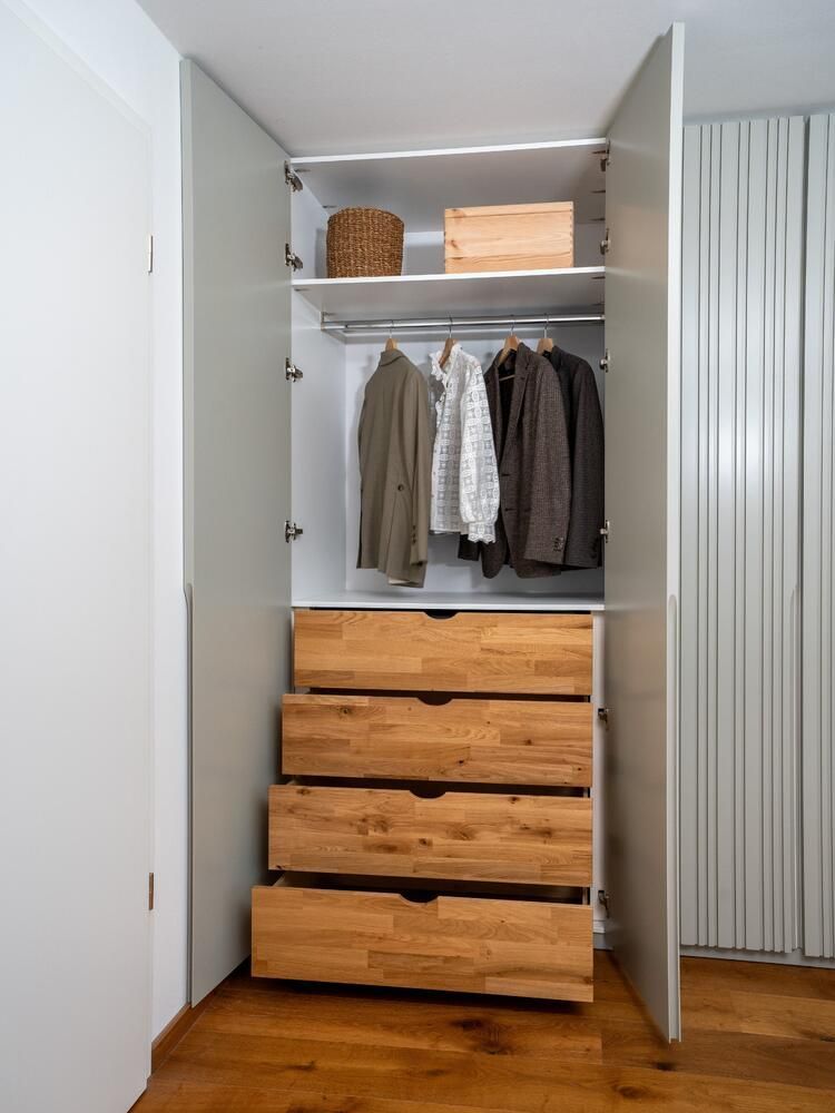 Offener Kleiderschrank mit grauen Türen, Holzschubladen und hängender Kleidung. Auf dem obersten Regal stehen ein Korb und eine Kiste.