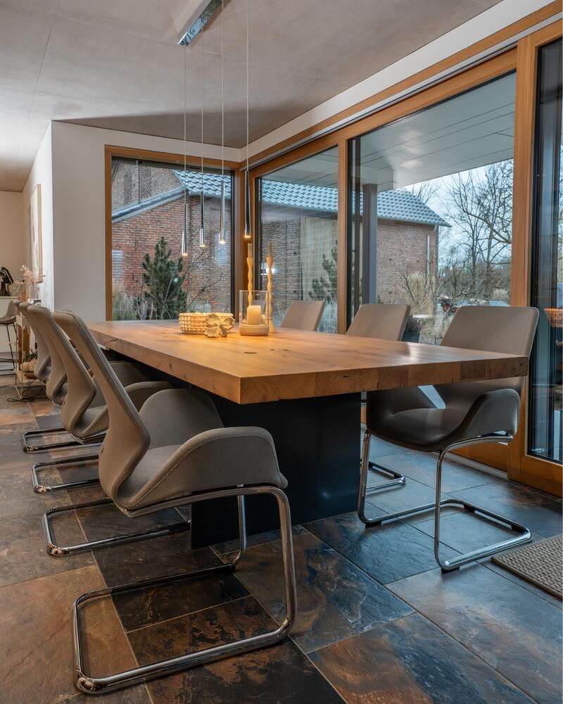Esszimmer mit großem Holztisch, grauen Stühlen und großen Fenstern mit Blick auf einen Hof.
