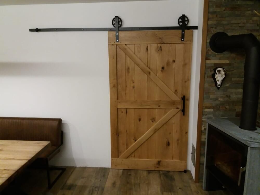 Eine hölzerne Scheunentür mit schwarzen Beschlägen, montiert an einer weißen Wand über einem Holzboden.