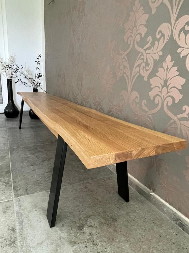 Eine Holzbank mit schwarzen Metallbeinen vor einer gemusterten Tapete und einer weißen Wand.
