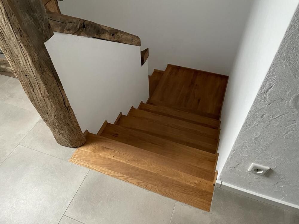 Eine Holztreppe führt vom Obergeschoss hinab. Links ist ein Balken sichtbar, die Wände sind weiß und der Boden grau gefliest.