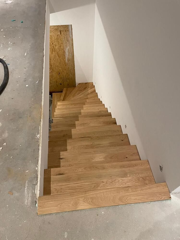 Eine Holztreppe in einem schmalen Flur führt hinunter zu einer Tür. Weiße Wände und Betonboden.