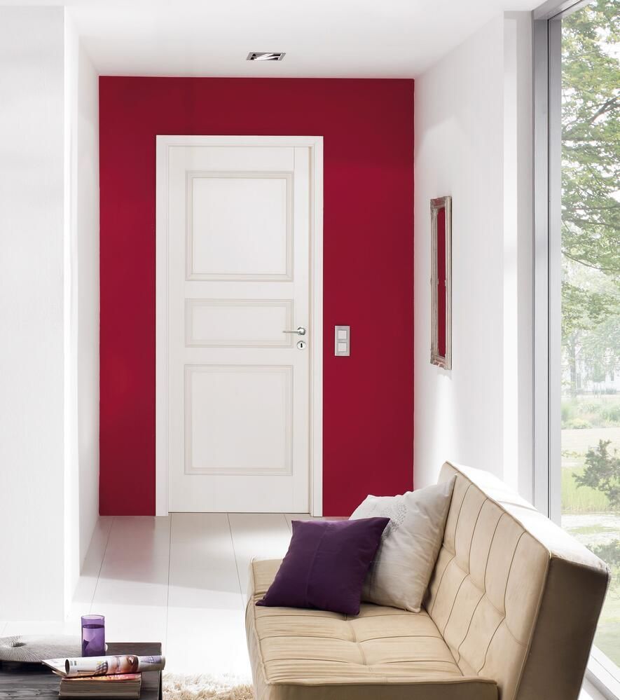 Eine weiße Tür, eingerahmt von einer roten Wand, führt in einen hellen Flur mit Sofa und Fenster.