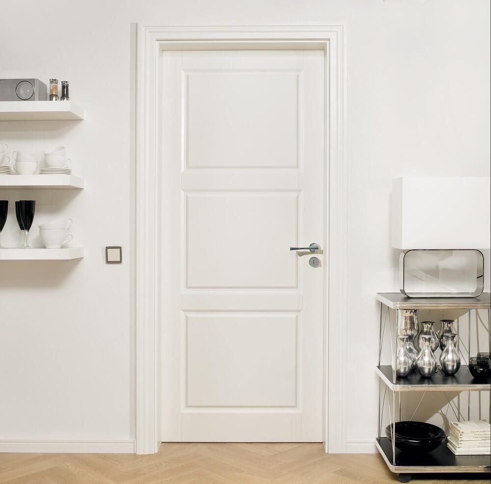 Weiße Paneeltür in einem weißen Raum. Links Regale mit Gegenständen, rechts ein Metalltisch.