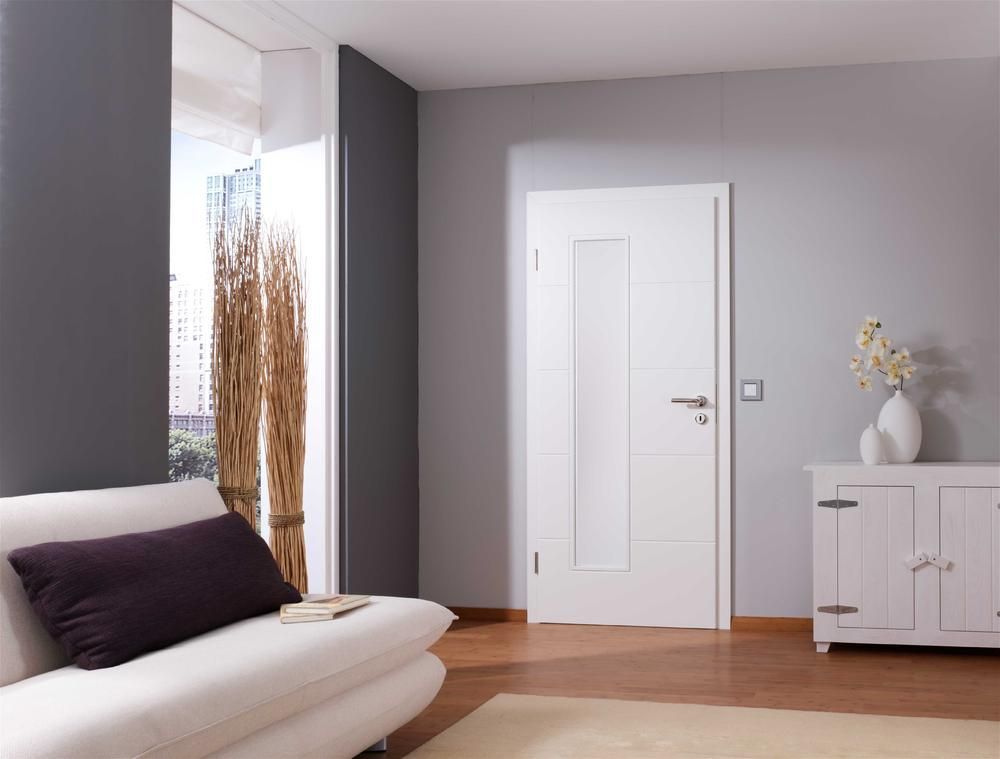 Weiße Tür in einem grau gestrichenen Zimmer mit einem weißen Sofa, einem Schrank und einem Fenster mit hohen, trockenen Pflanzen.
