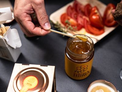 Eine Hand rührt Honig in einem Glas neben aufgeschnittenen Tomaten auf einem dunklen Tisch