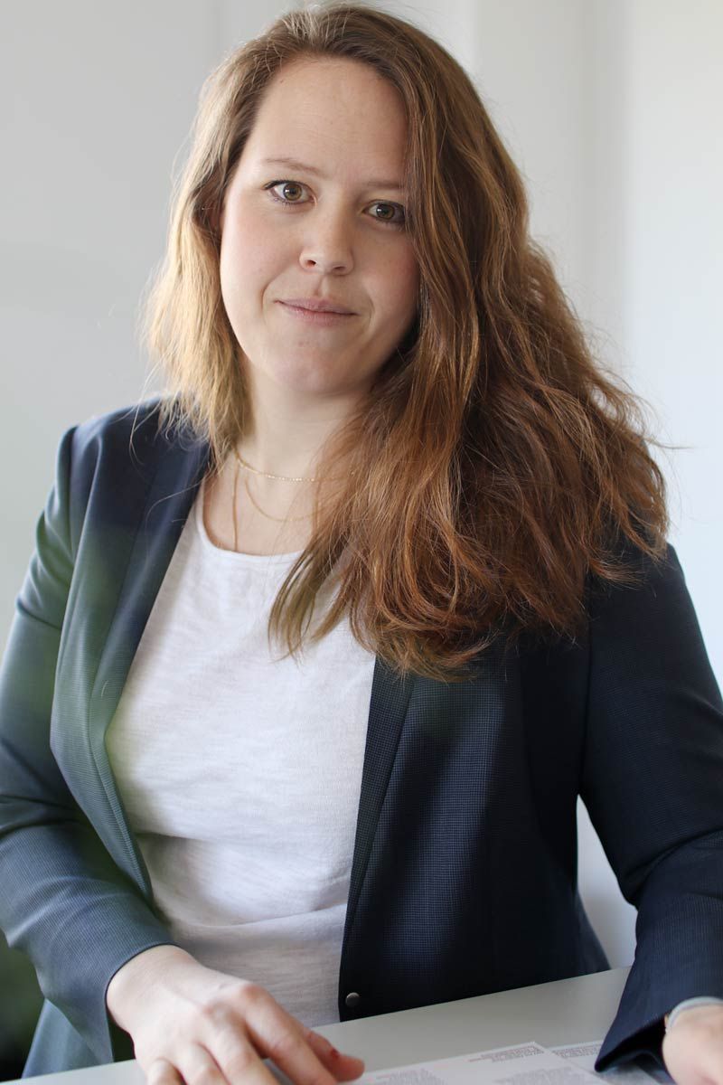 Frau in Blazer und weißem Hemd, die in die Kamera schaut, mit langen braunen Haaren, sitzt an einem Schreibtisch.