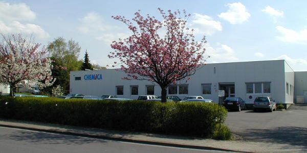 Weißes Gebäude mit blauem Logo, geparkte Autos und blühende Bäume.