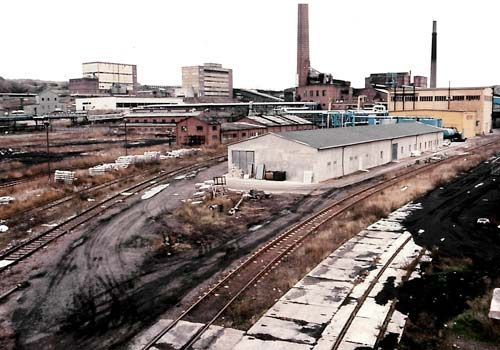 Industriegelände mit Bahngleisen, Gebäuden, Schornsteinen und einem Lagergebäude.