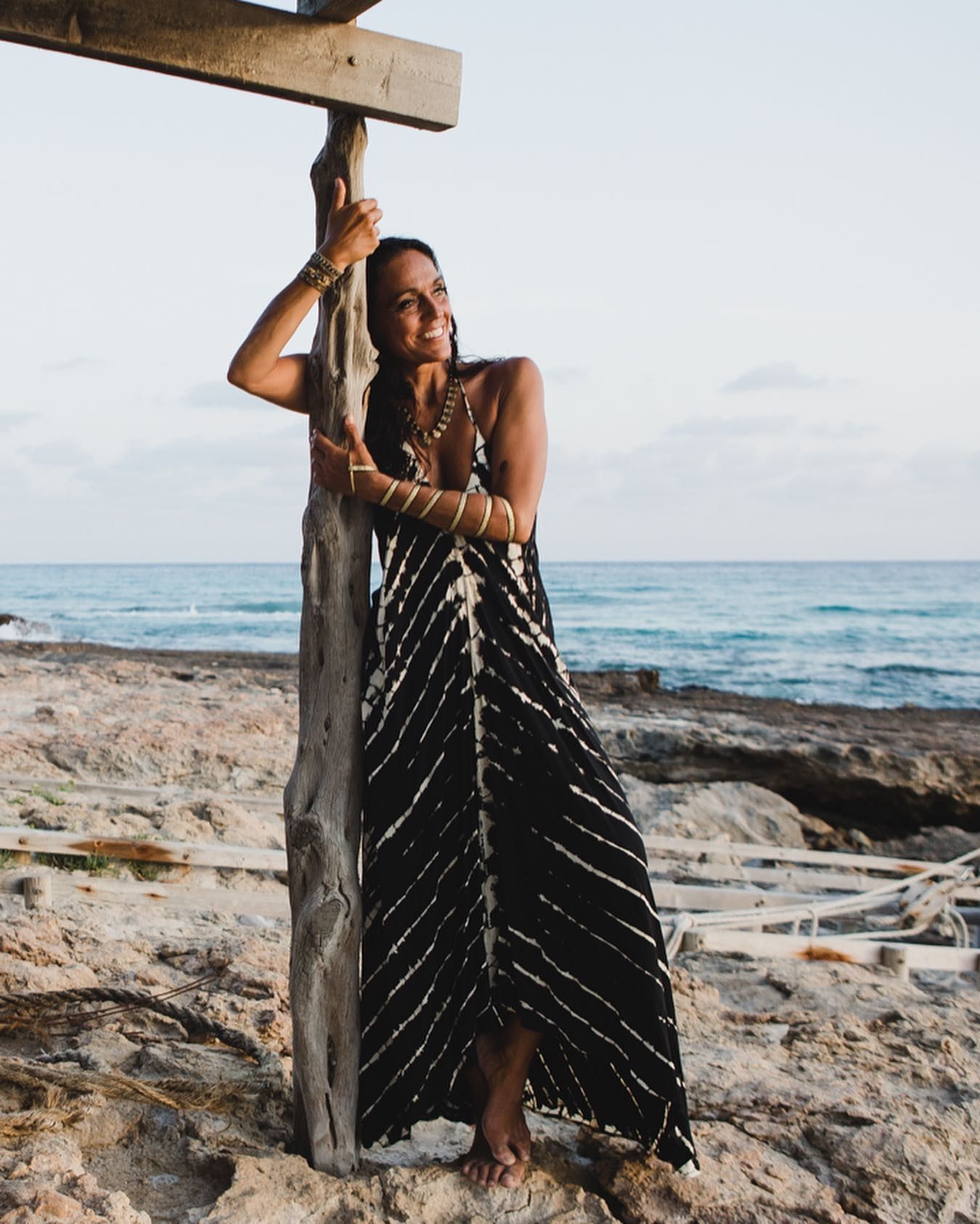 Mujer con un vestido de rayas negras apoyada en un poste de madera en una playa de arena junto al océano.