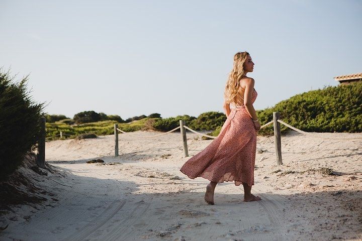 Mujer con vestido rosa caminando por un sendero arenoso cerca de dunas y vegetación bajo un cielo brillante.