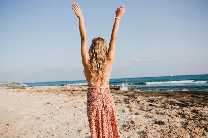 Persona con los brazos en alto en una playa de arena junto al océano, vestida con un vestido rosa.