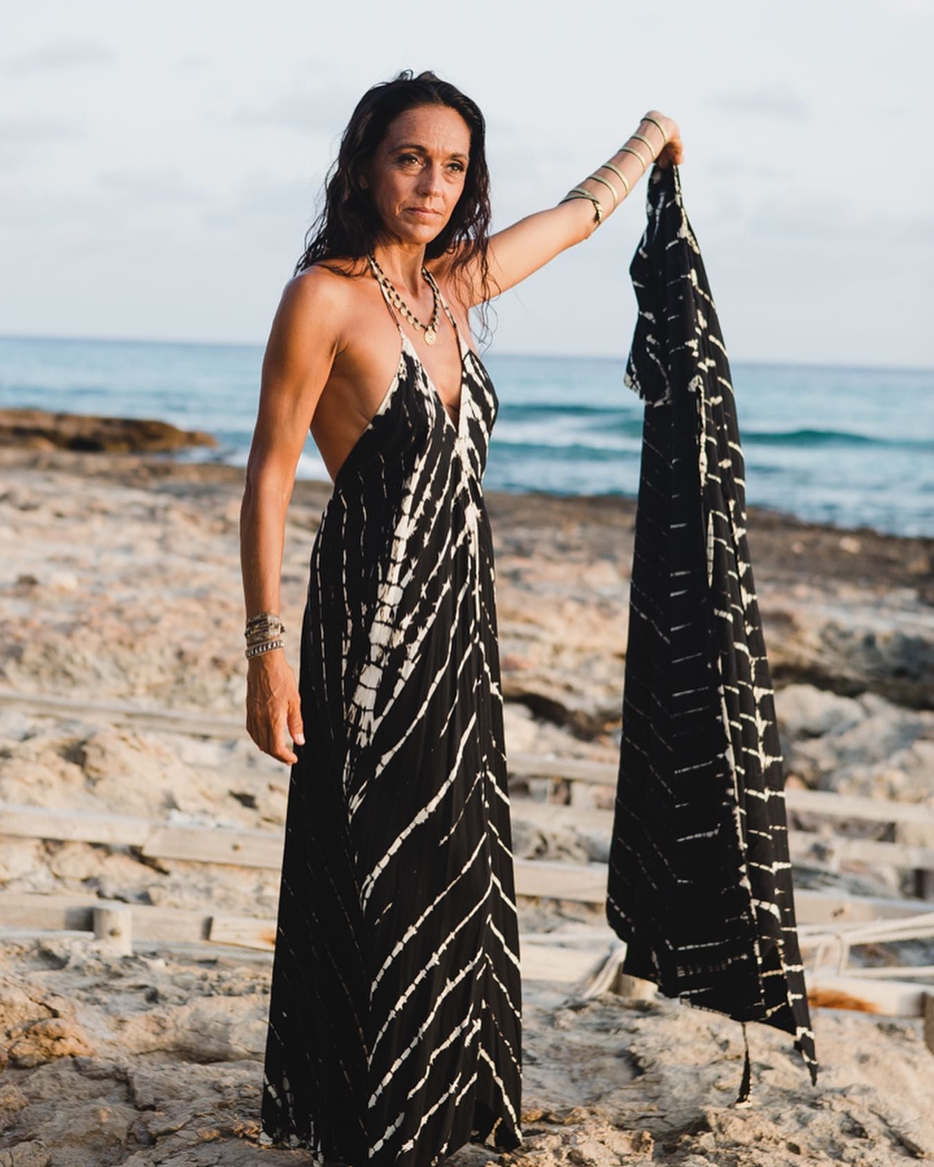 Mujer con un vestido blanco y negro de pie en una playa rocosa, sosteniendo la tela del vestido.