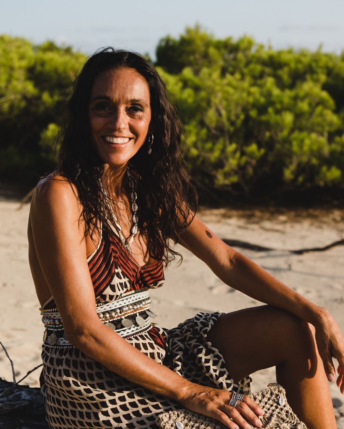 Una mujer sonríe en la playa, sentada en la arena, con un vestido estampado y arbustos verdes detrás de ella.