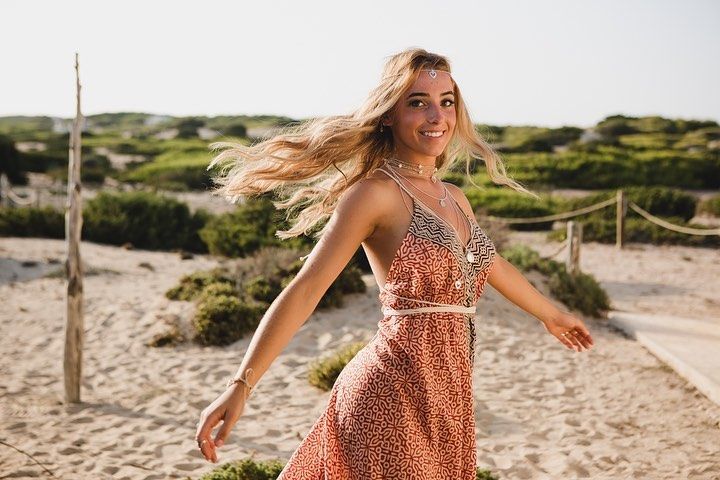 Mujer con un vestido estampado caminando por una playa de arena con dunas al fondo.