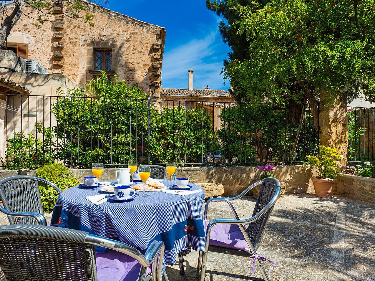 Outdoor cafรฉ table with blue checkered cloth in a sunny stone courtyard, surrounded by greenery
