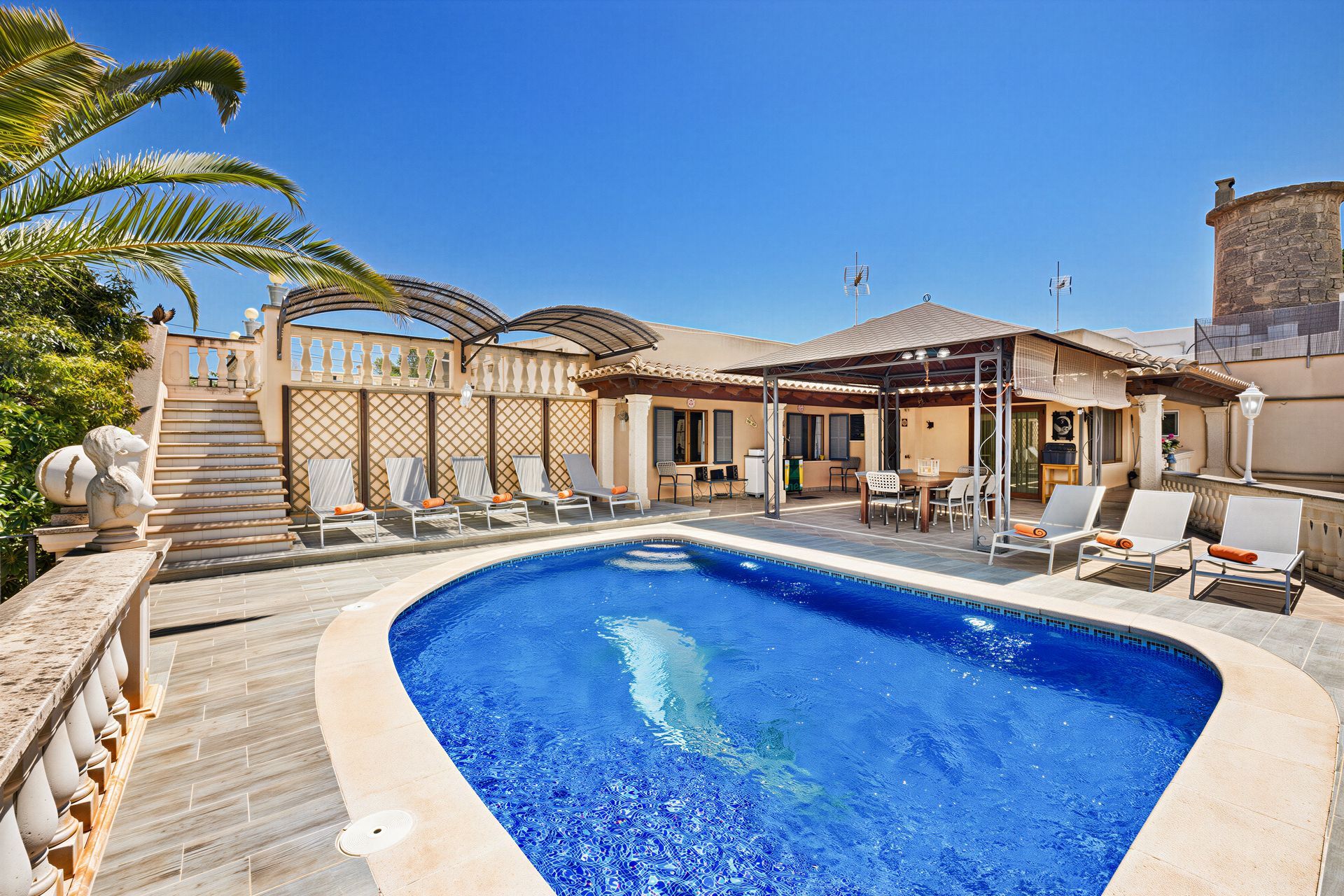 Sunny patio with a blue swimming pool, lounge chairs, palm trees, and a villa-style house.