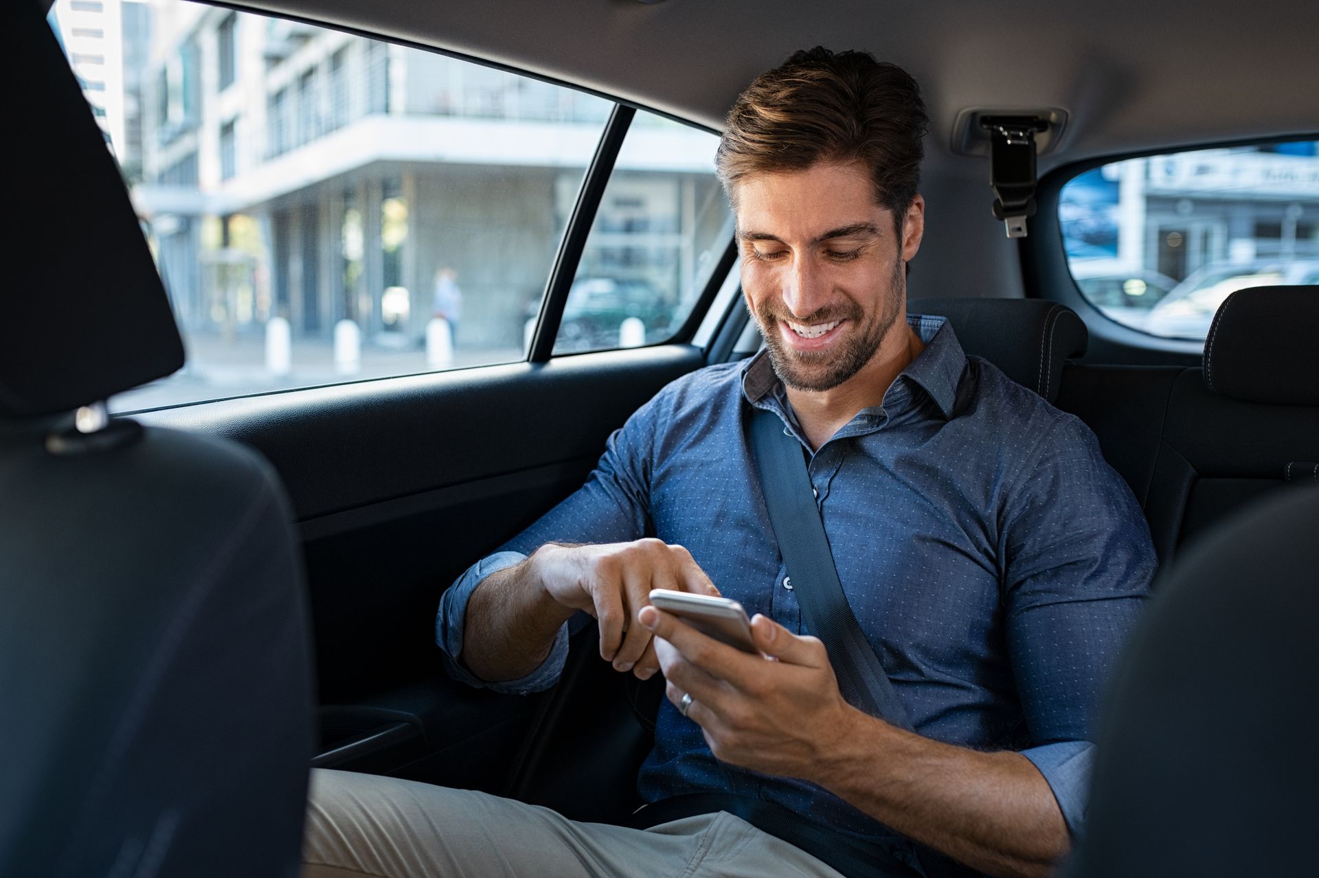 Un homme en chemise utilise son téléphone, souriant, assis à l'arrière d'une voiture.