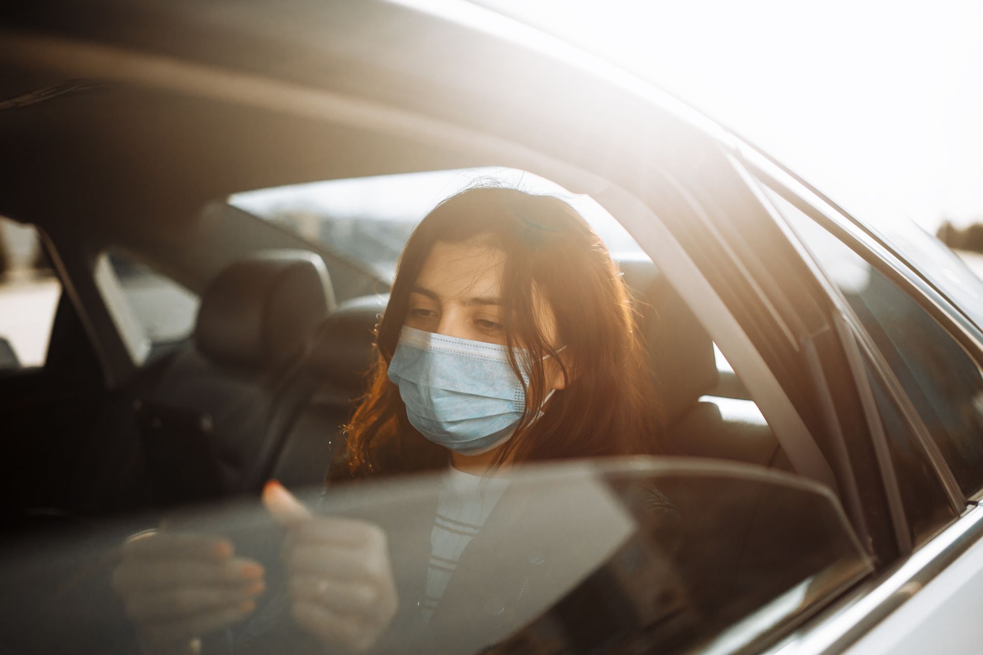 Une femme portant un masque est assise à l'arrière d'une voiture, le regard baissé. La lumière du soleil filtre à travers la vitre.