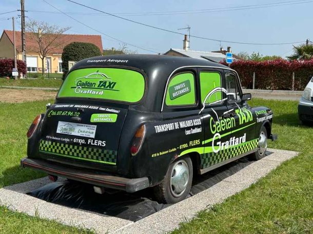 Un taxi de style londonien, arborant le logo vert Gaetan Taxi Graffard, est stationné sur une dalle de béton.