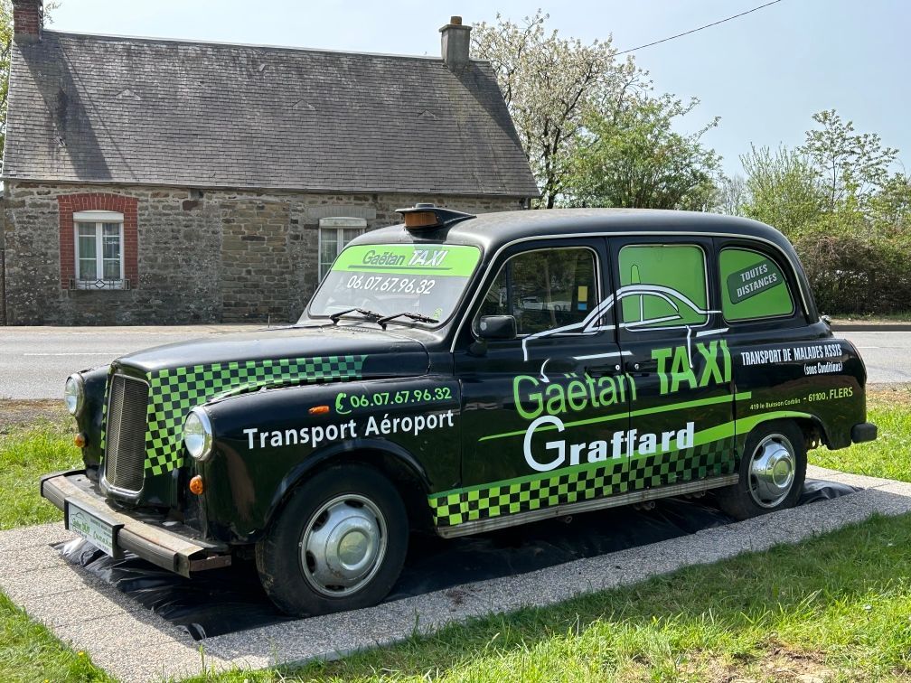 Un taxi londonien à carreaux, portant l'inscription Gaëtan Graffard Taxi, est garé sur du gravier devant une maison en pierre.