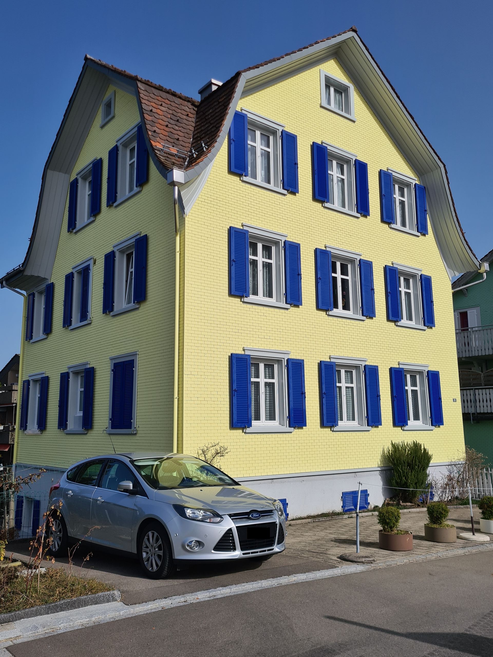 Gelbes dreistöckiges Haus mit blauen Fensterläden, rotem Dach und einem silbernen Auto davor.