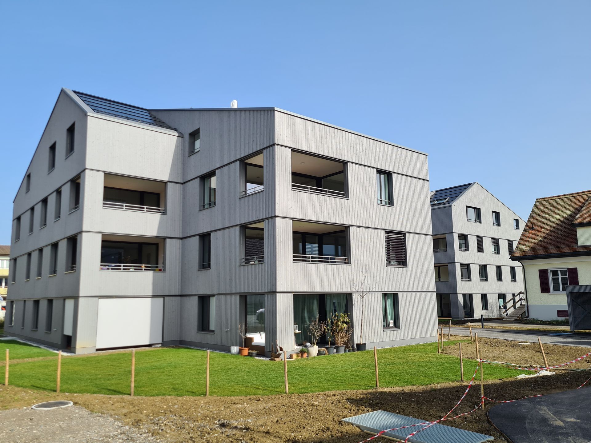 Graue, moderne Apartmentgebäude mit Balkonen, eingebettet in eine Rasenfläche an einem sonnigen Tag.