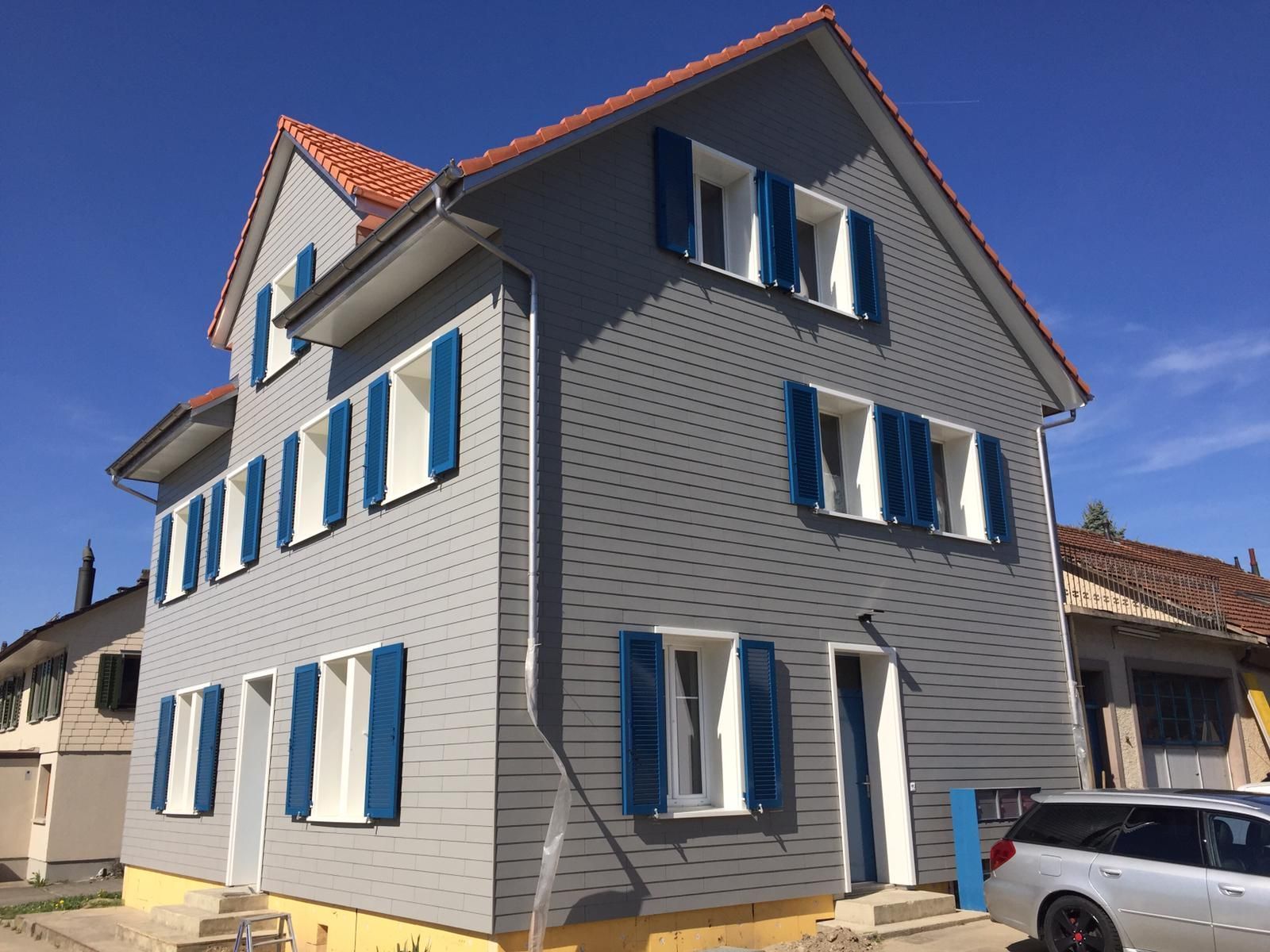 Graues Haus mit blauen Fensterläden und orangefarbenem Dach.