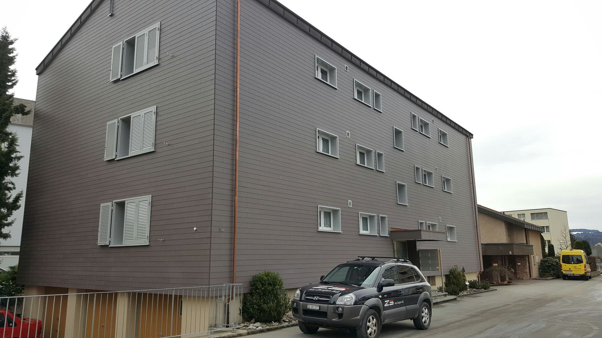 Dreistöckiges braunes Gebäude mit mehreren Fenstern. Davor parkt ein dunkler Geländewagen.