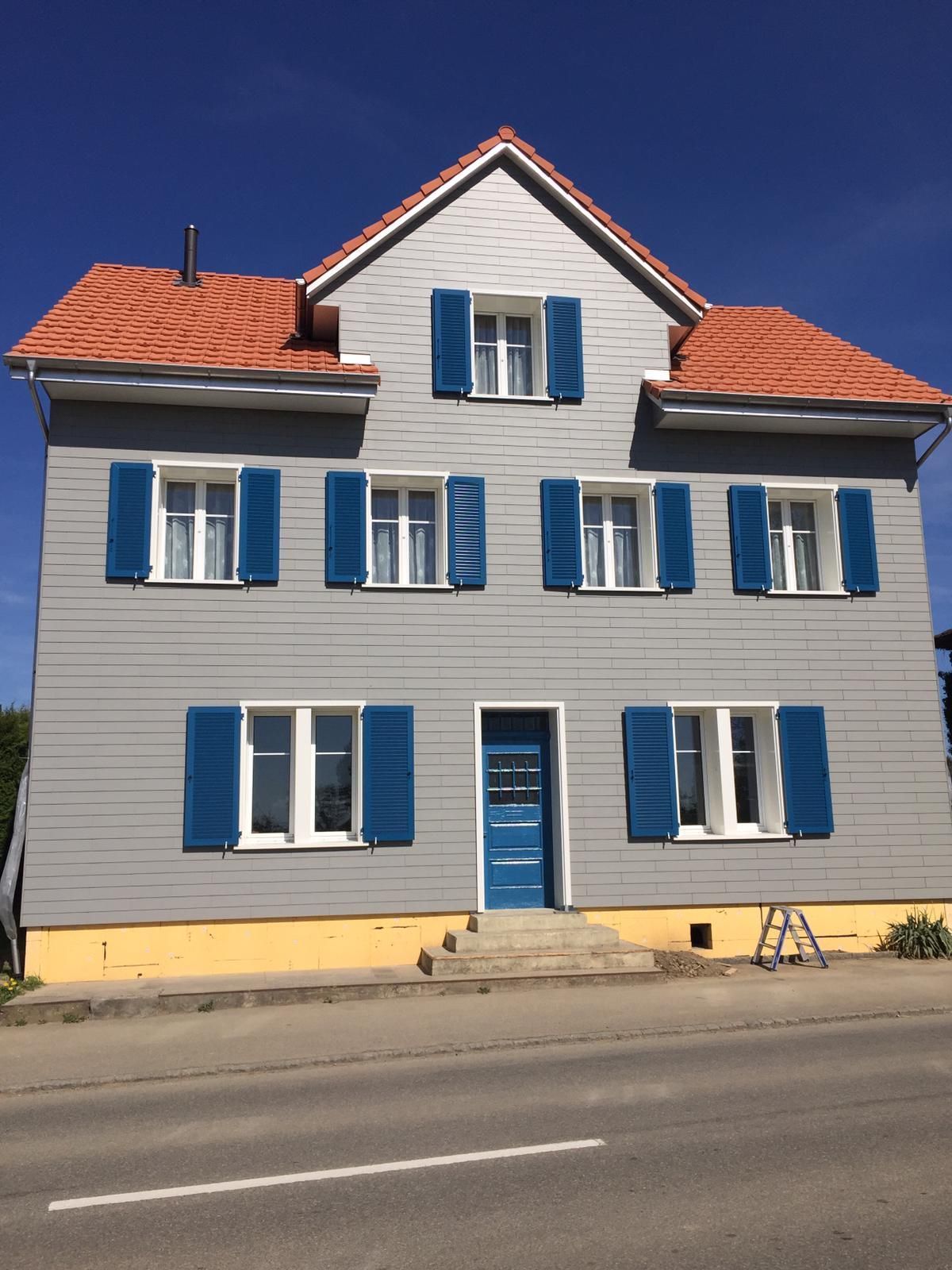 Dreistöckiges Haus mit grauer Fassade, blauen Fensterläden und Tür, rotem Ziegeldach und blauem Himmel.