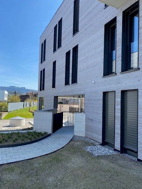 Gemütliches Wohnzimmer mit grauem Sofa, Couchtisch aus Holz, Backsteinwand und grossen Fenstern mit Pflanzen.