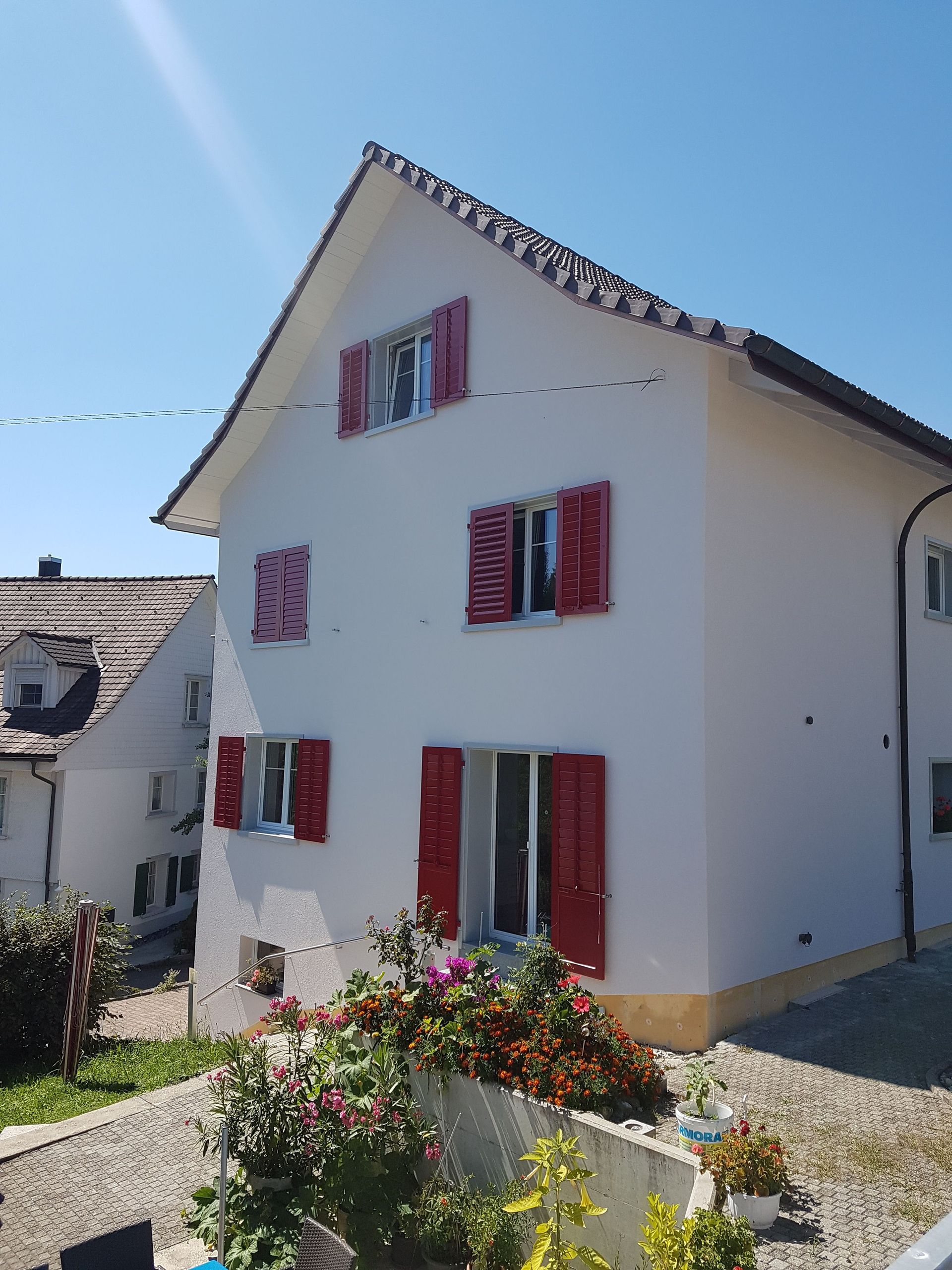 Weisses Haus mit roten Fensterläden, blühenden Sträuchern und blauem Himmel.