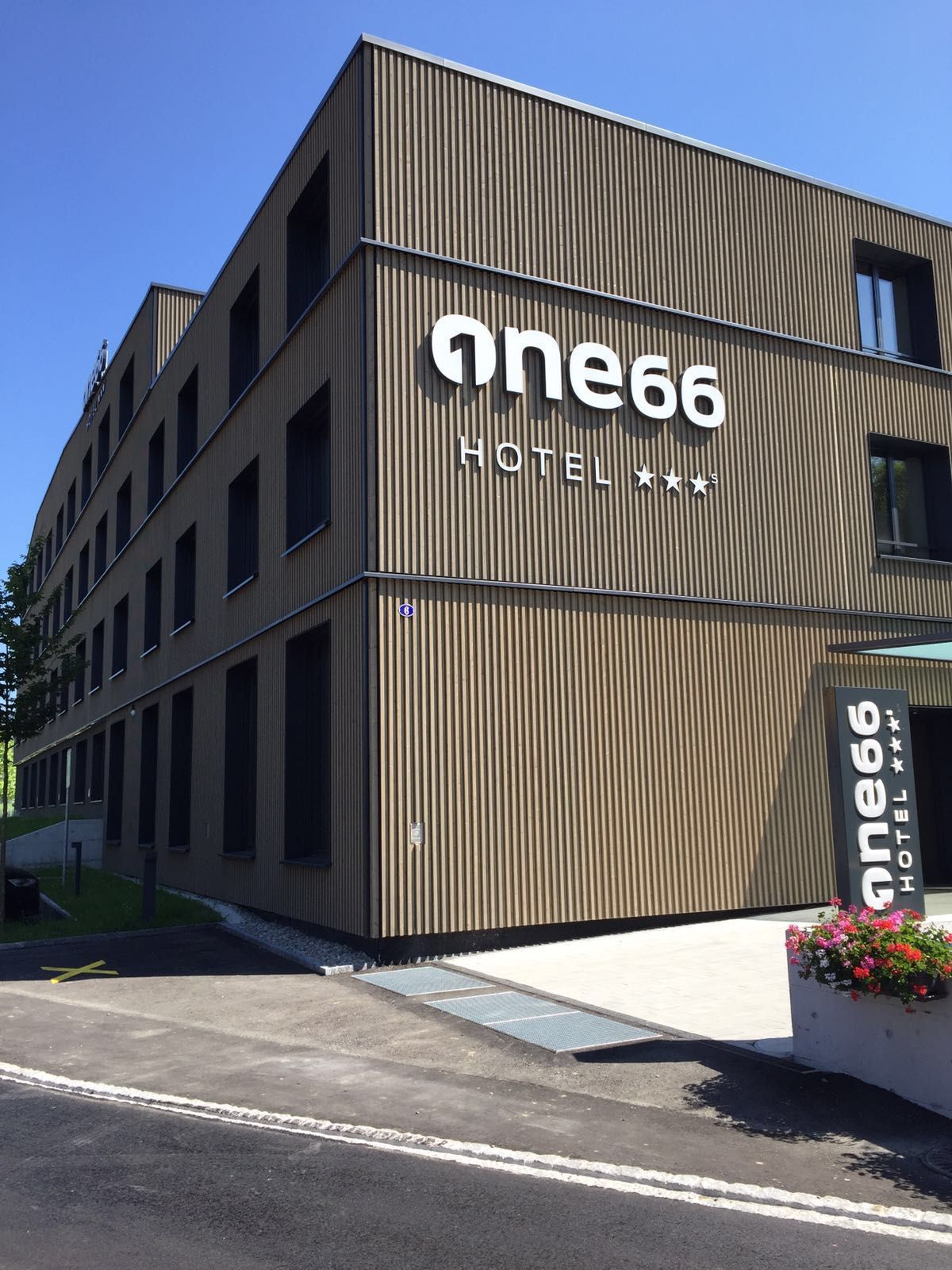 Modernes Gebäude mit dem Schild „one66 HOTEL ***“. Braune Holzfassade, Fenster, sonniger Tag.