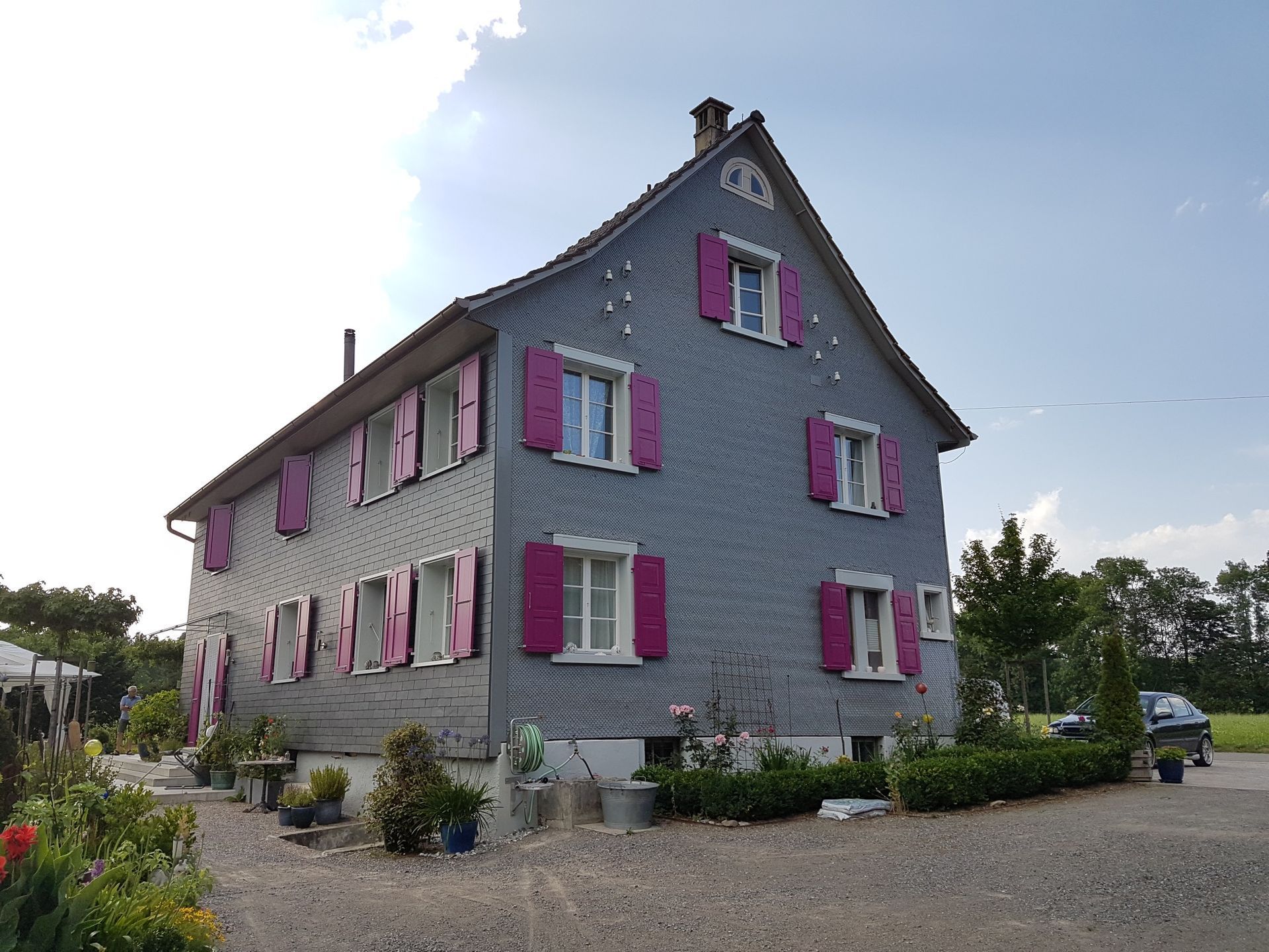 Zweistöckiges Haus mit grauer Fassade, rosa Fensterläden und weissen Fensterrahmen. Es liegt in einer Rasenfläche mit einer Kiesauffahrt.