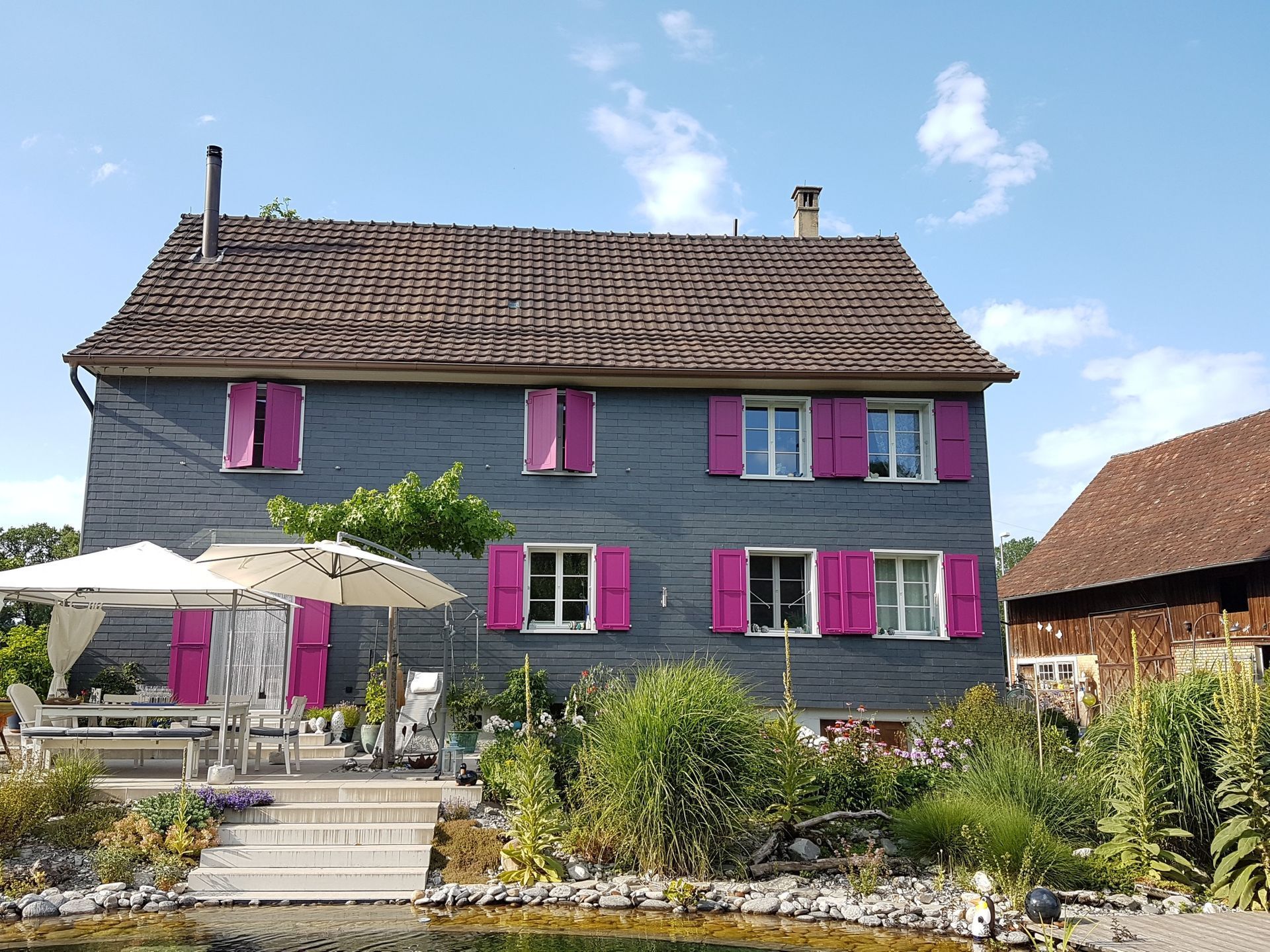 Zweistöckiges graues Haus mit rosa Fensterläden. Weisser Essbereich im Freien. Braunes Dach am Haus und an einem Gebäude rechts davon.