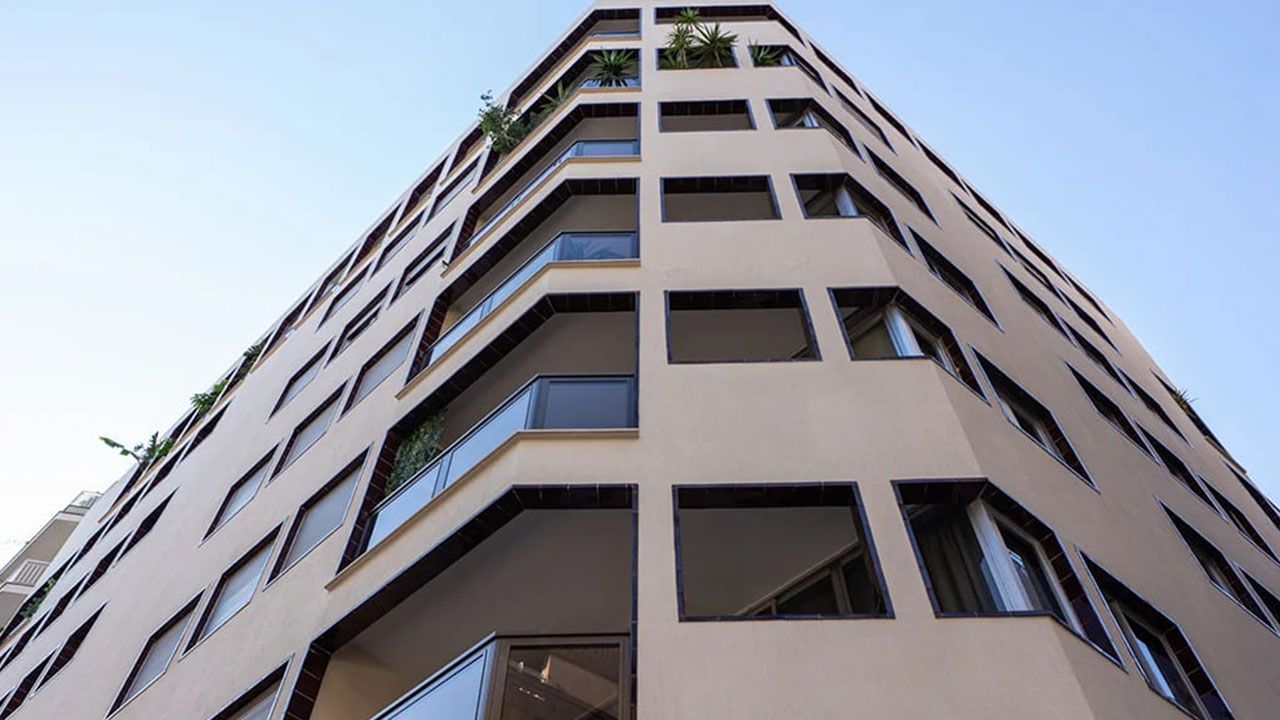 Vista desde un ángulo bajo de un edificio beige de varias plantas con ventanas oscuras contra un cielo azul.