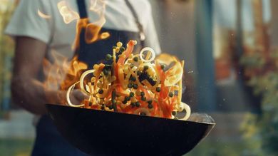 A man is cooking vegetables in a wok with flames coming out of it.