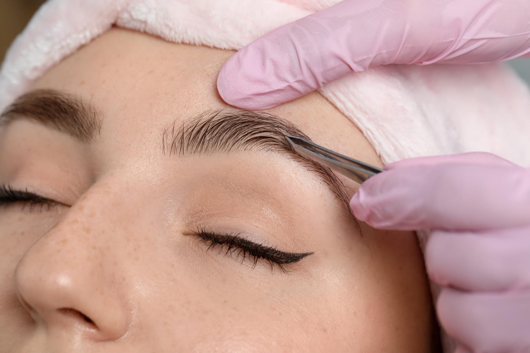 Primer plano de la depilación de cejas con pinzas sobre el ojo cerrado de una persona.