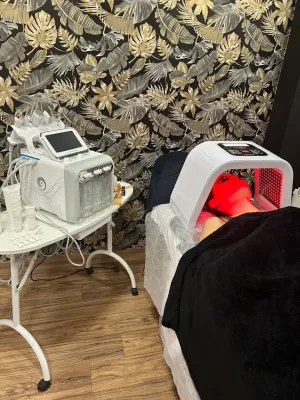 Sala de spa con un aparato de terapia facial iluminado en rojo junto a una camilla de tratamiento y un carrito con ruedas.
