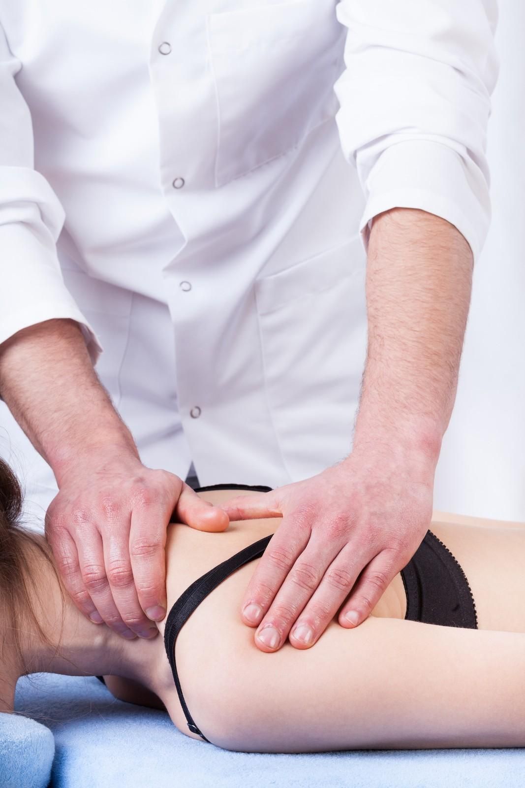 Masajista trabajando el hombro y el cuello de una persona. La persona está tumbada y lleva una camiseta negra.