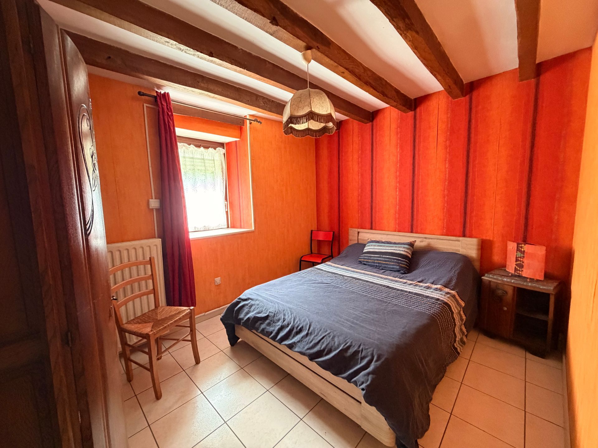 Petite chambre aux murs orange, poutres en bois, un lit, une chaise et une fenêtre avec des rideaux rouges.