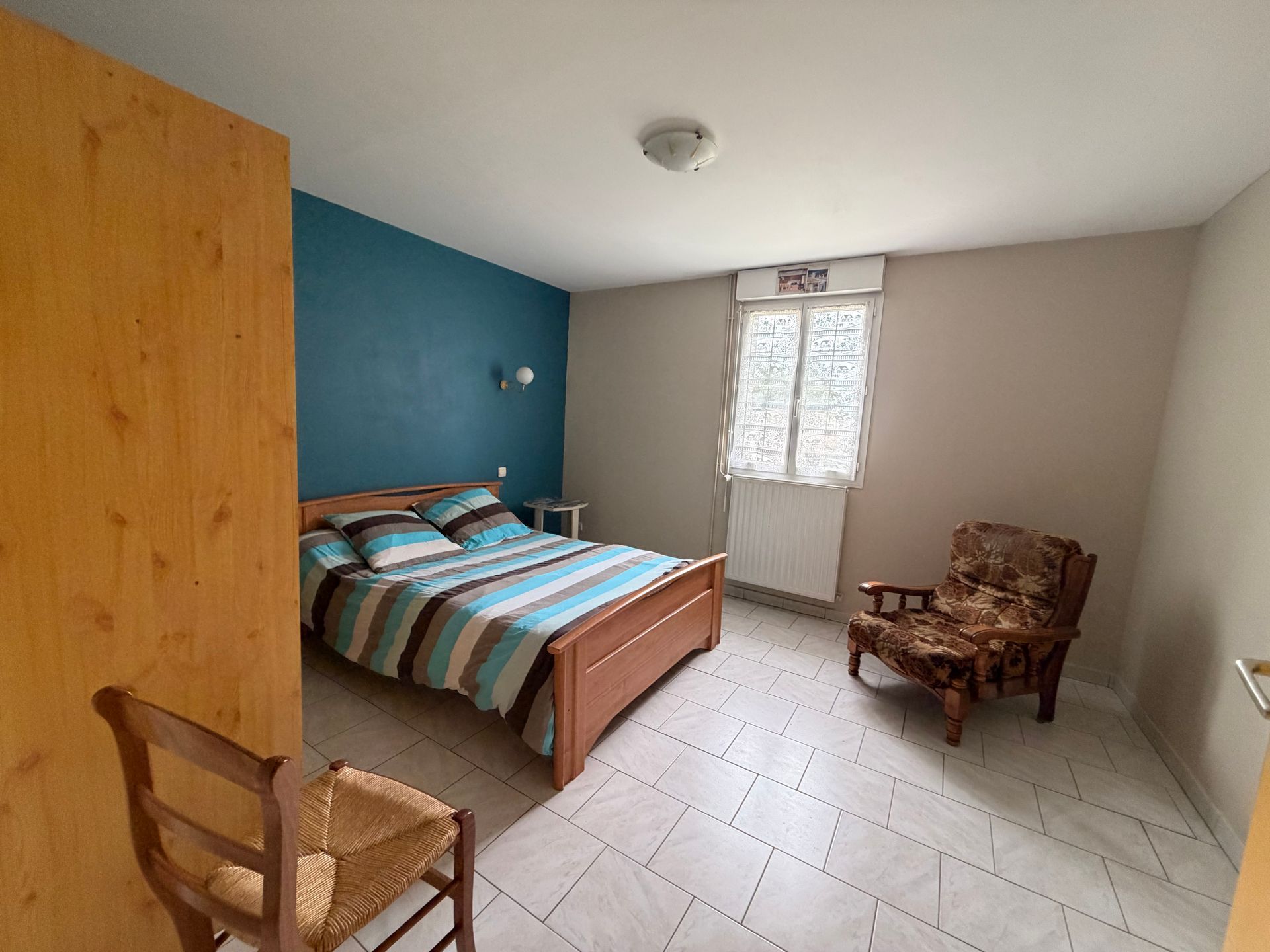 Chambre d'amis avec un lit, un mur d'accent turquoise, une fenêtre, un fauteuil en osier et un sol carrelé.
